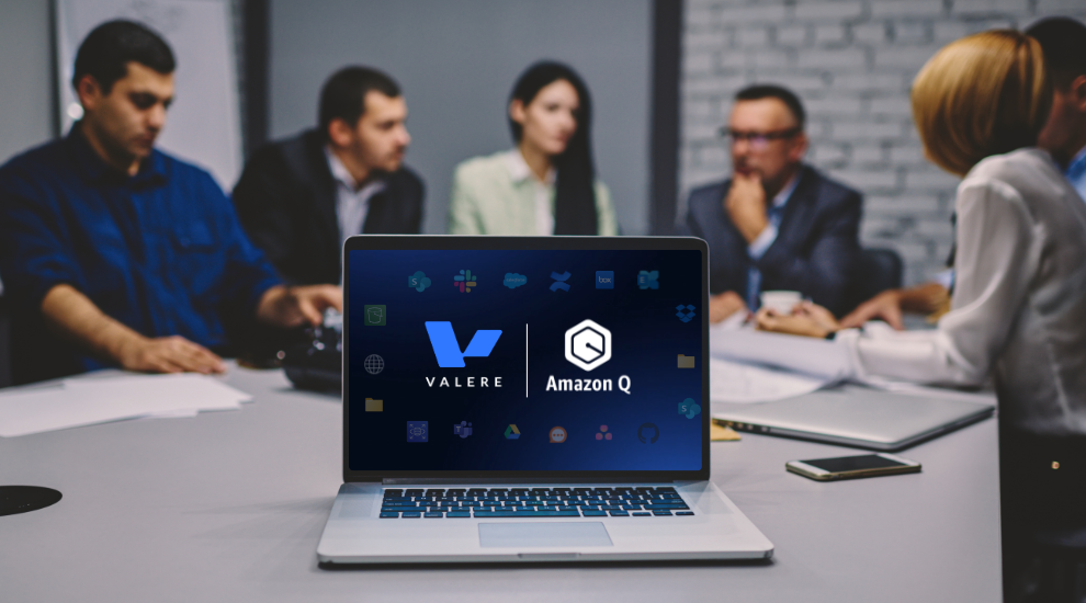 A laptop on a conference table displays the Valere and Amazon Q logos. In the blurred background, several people are seated around the table in a meeting.