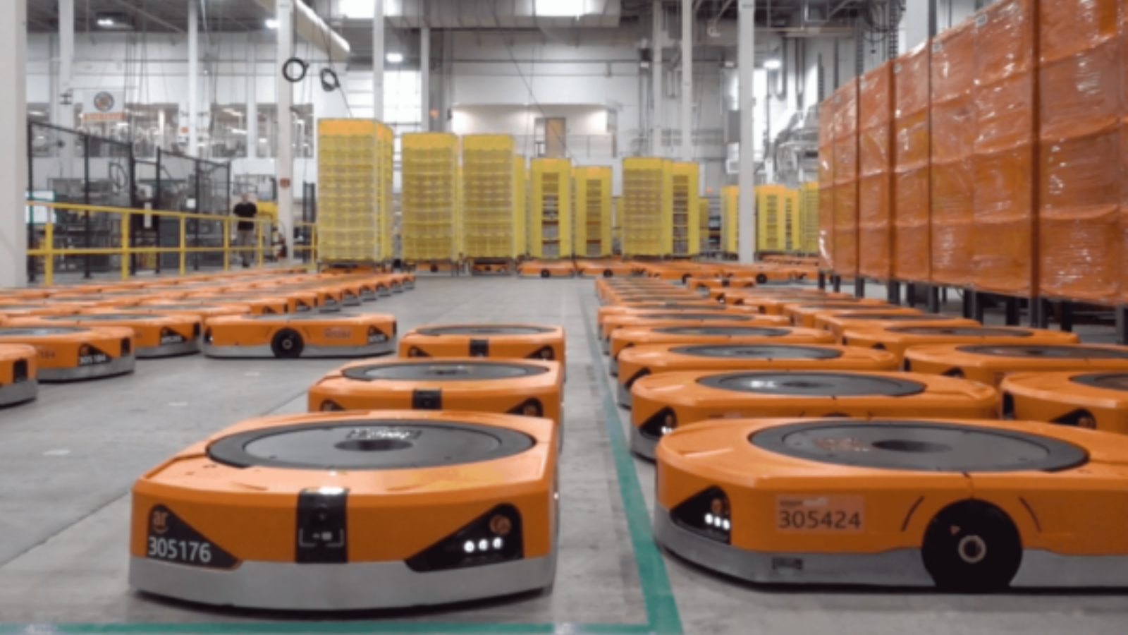 Warehouse floor with multiple autonomous robots transporting shelves filled with packages—showcasing Amazon’s robotic logistics system in action.